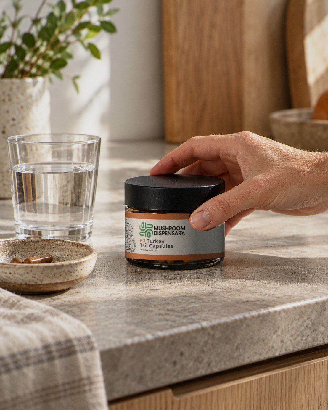 A hand reaching for a jar of Mushroom Dispensary Turkey Tail Capsules on a kitchen counter beside a glass of water